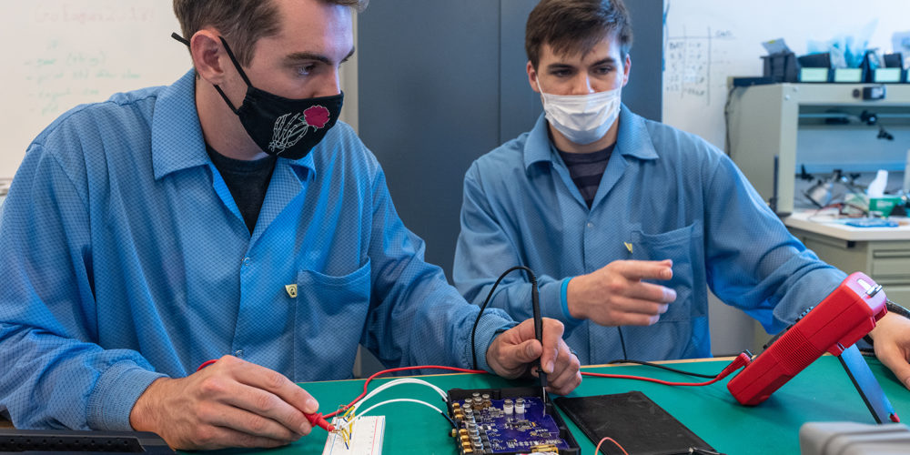 Two students in lab coats work on an electronic panel