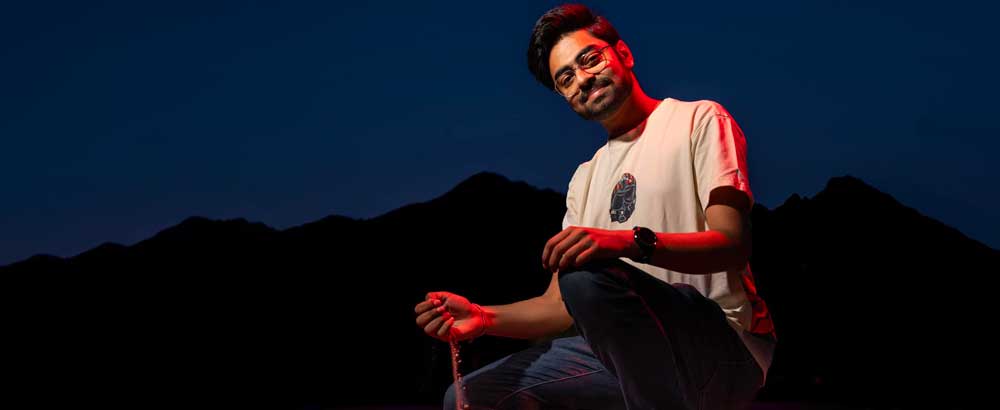 Aashman Gupta squats with sand sifting though his fingers in front of mountain range