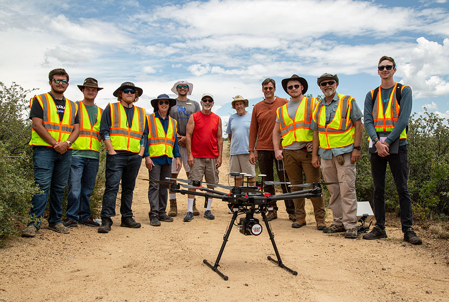 A group of students and faculty wearing high-visiblity vests, near a multirotor weather research drone