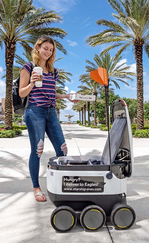 Woman holding a pizza box and cup of coffee next to an open food delivery robot