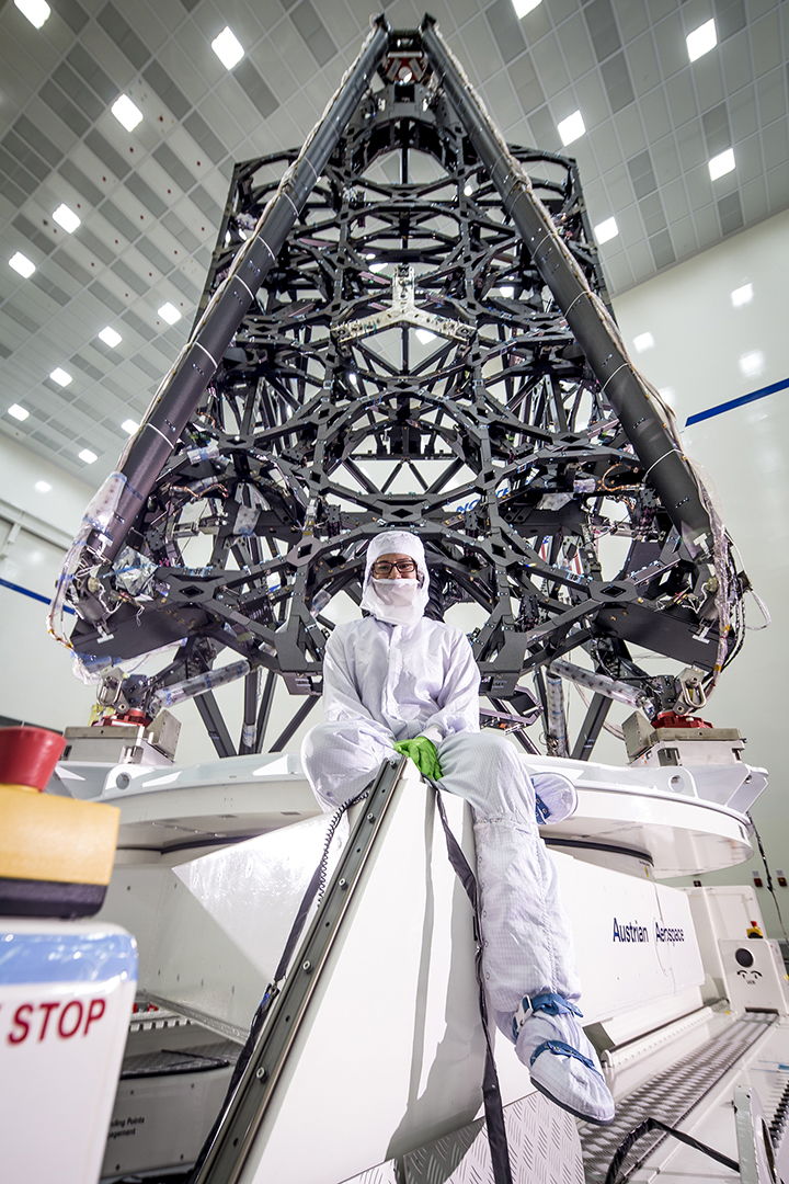 Tasha Graciano wears a clean room suit and sits in front of the skeleton of the James Webb Space Telescope.