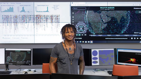 Moriba Jah stands in front of several computer monitors and projection screens.