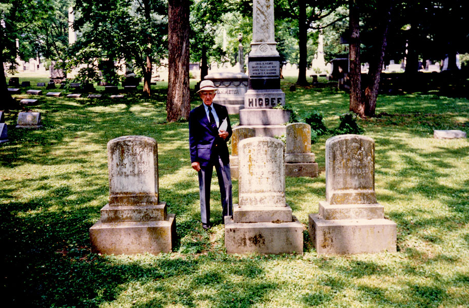 John Paul Riddle standing in a cemetery next to T. Higbee Embry's gravestone.