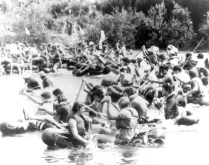 black and white photo of a large group of men on inflatable boats paddling hard in every direction