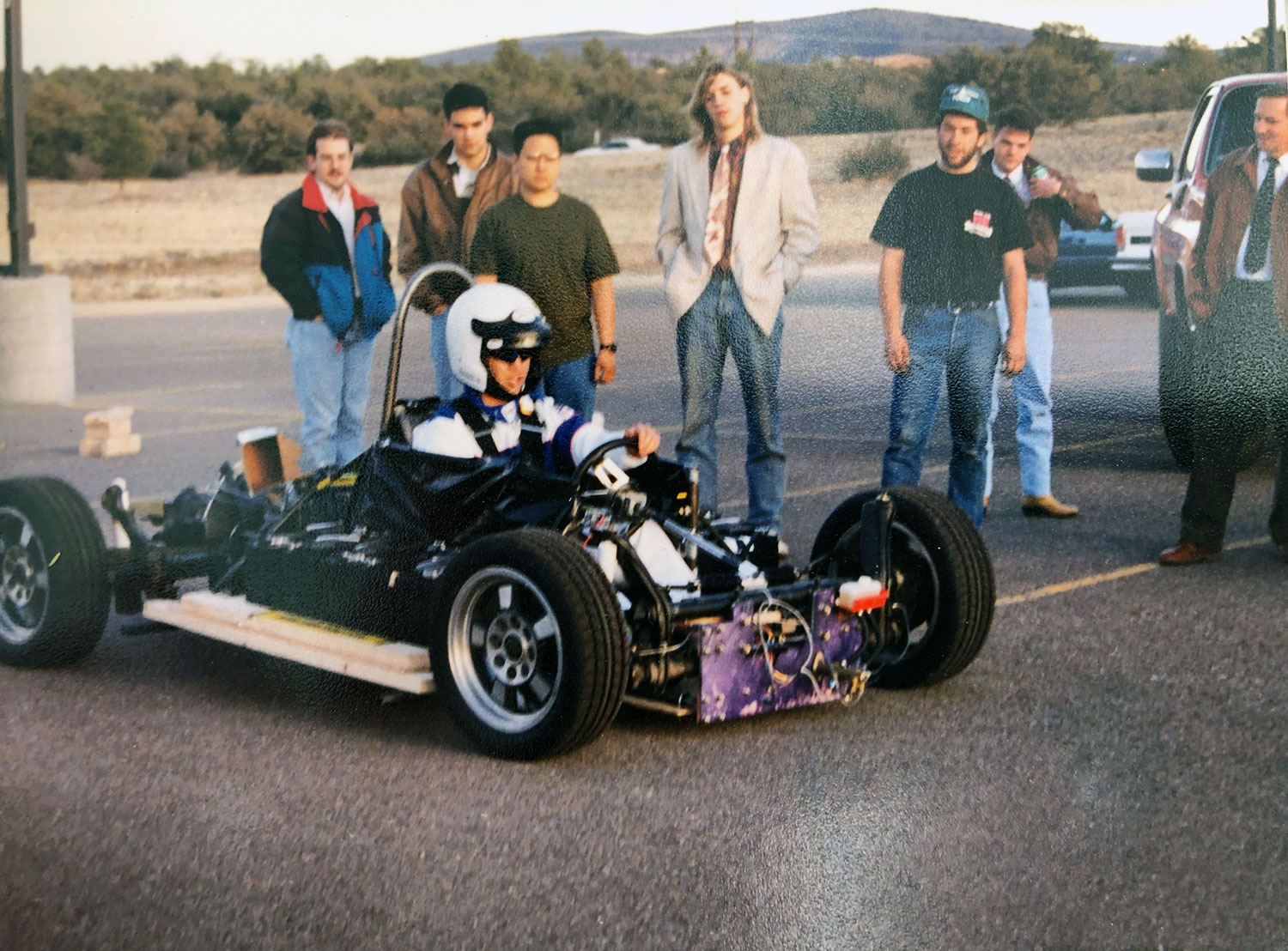 An electric car chassis in a parking lot with driver seated, other students standing nearby