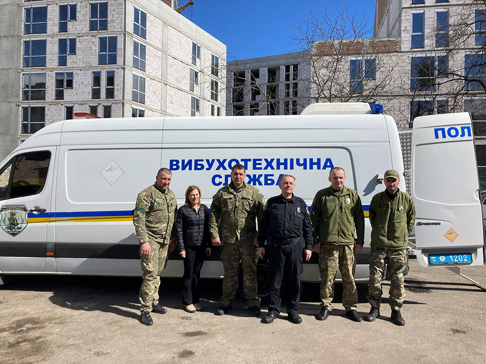 Oksana Bardygula stands with four men in uniform in front of a cargo van.